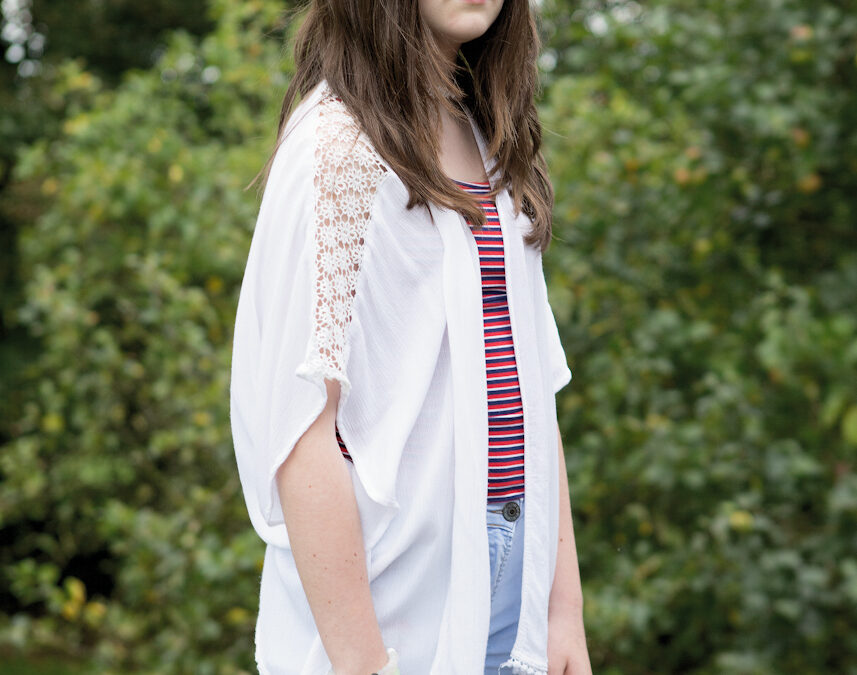 A portrait of a young woman, standing slightly to the side, looking at the camera, with greenery and planting in the background.
