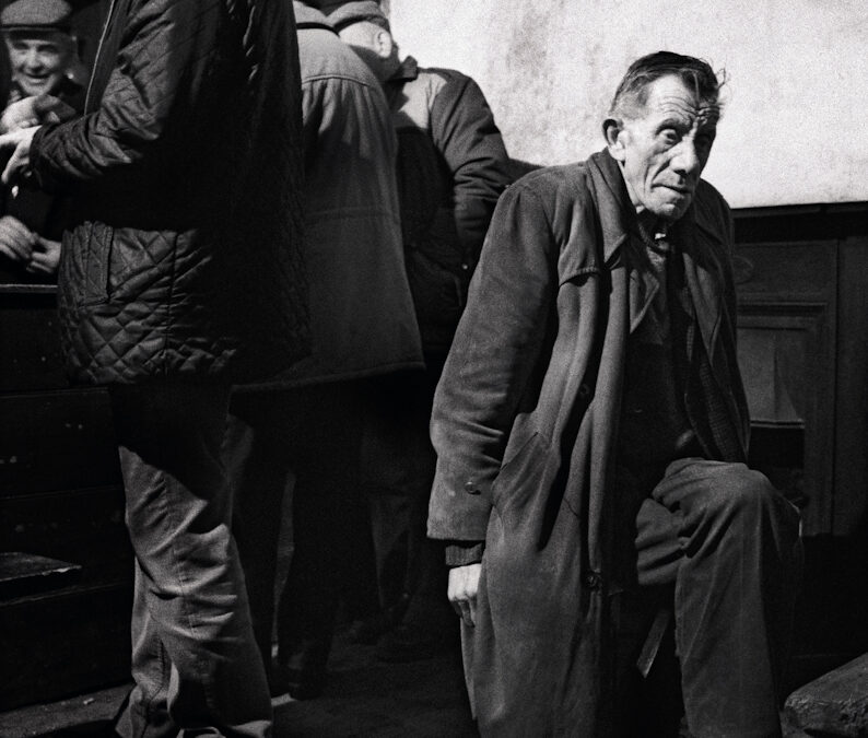 A black and white image. A man sits in a long coat, behind him a number of men lean against the bar in a pub.