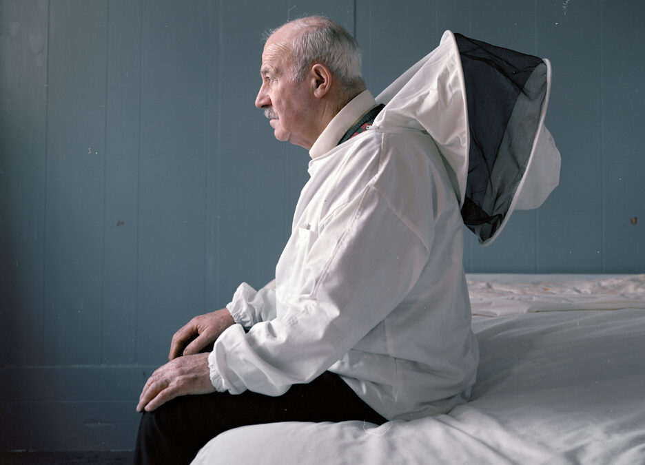 An older man sits on the edge of a seat covered with white linen. He is dressed in a white beekeepers outfit.