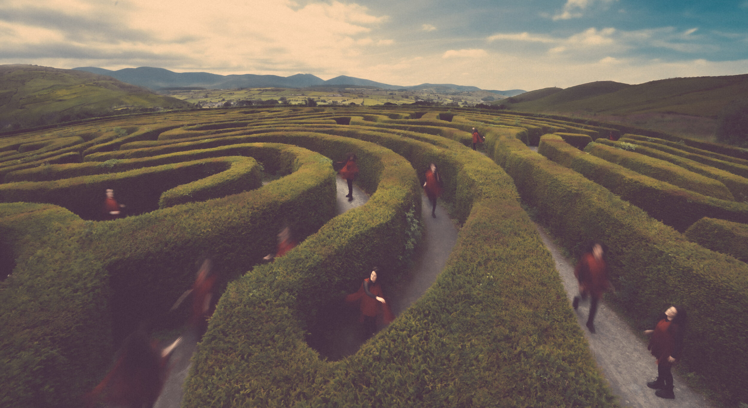 A maze with tall green bushes from a view from above that shows a woman in red clothes multiple times in differents parts of the maze as she tries to complete the maze