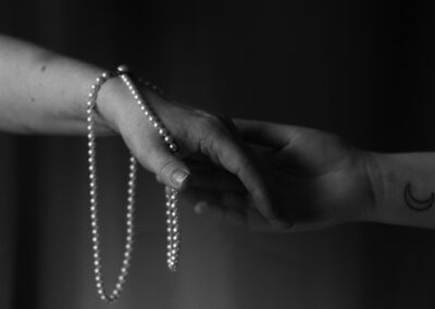 A black and white photograph showing two hands gently touching. One hand is adorned with a strand of pearls draped over it, while the other hand has a crescent moon tattoo on the wrist. The image captures a sense of connection and intimacy against a dark, blurred background.