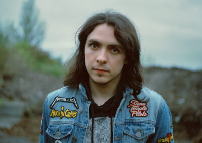 A portrait of a young person with long dark hair, standing outdoors against a blurred, natural background. They are wearing a blue denim jacket adorned with patches of rock bands like Metallica, Alice in Chains, Stone Temple Pilots, and Iron Maiden, among others. The expression on their face is calm and slightly introspective, and the overcast sky contributes to a subdued, moody atmosphere.