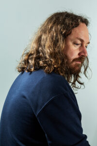 Artist Shane Hynan site with his back toward the camera. we see his face in a side profile. he has long hair, a beard, and is wearing a navy top.