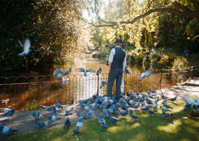 A man stands feeding pigeons by a pond surrounded by trees