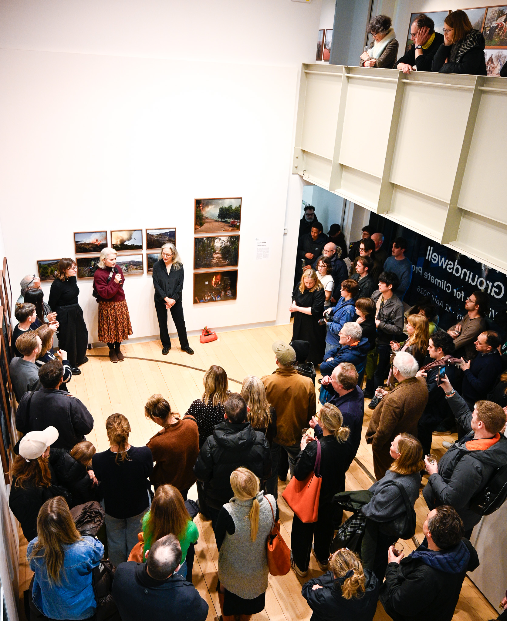 A young artist resident at Photo Museum Ireland discusses their work with a curator at a mentorig session. They are sat at a wooden table with a laptop. Behind them is a large tv screen showing their photographic work. In the foreground a notepad is visible.