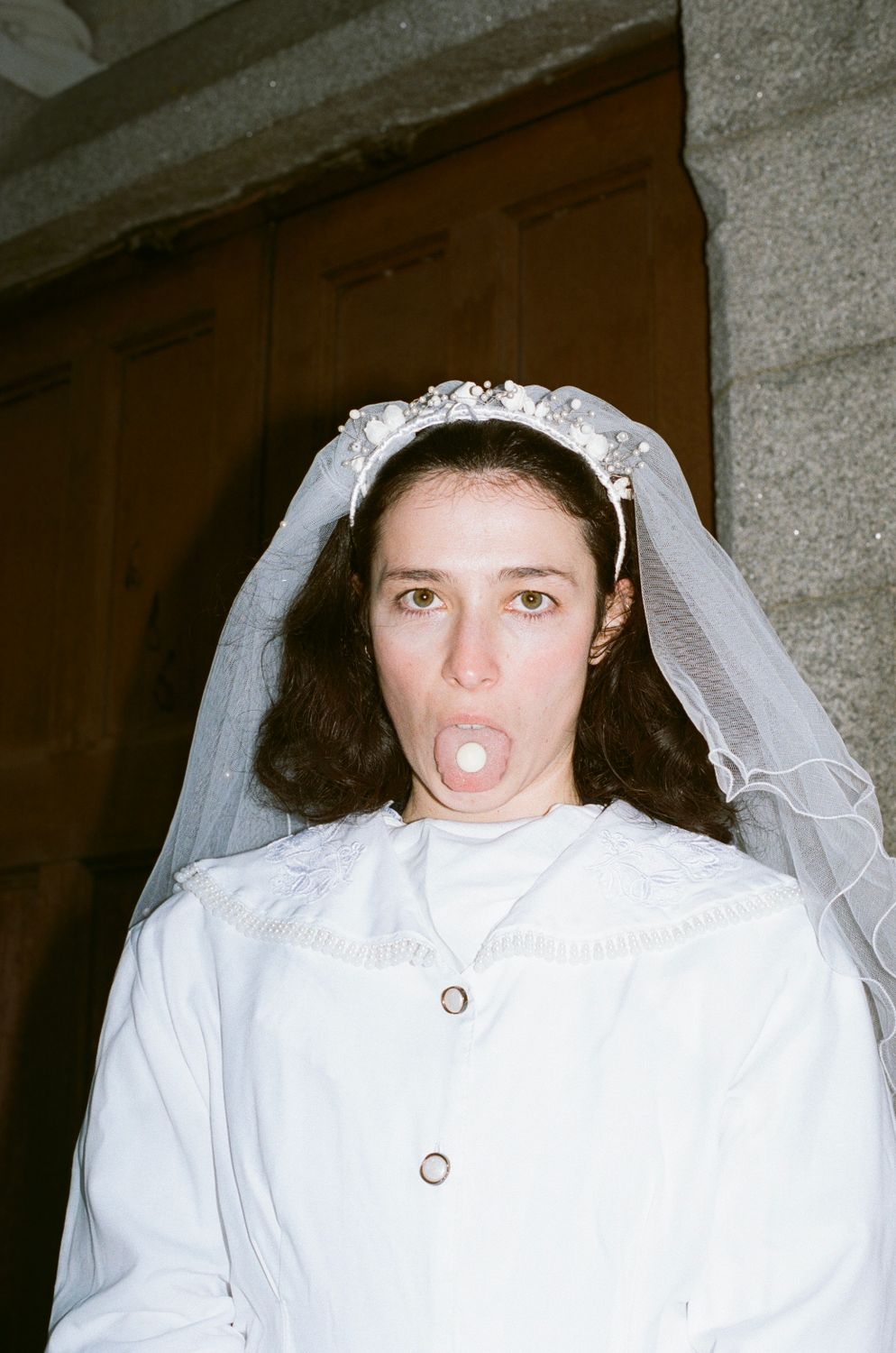 The image shows a person wearing a white garment with a high collar and button details. The garment has embroidered patters near the collar. The individual has shoulder length brown hair slightly curly and is also wearing a white veil with a decorative headpiece that includes floral elements and beads. The background features a stone wall and a wooden door. The person is looking straight ahead and their tongue is sticking out with what appears to be something small, round and white on their tongue.