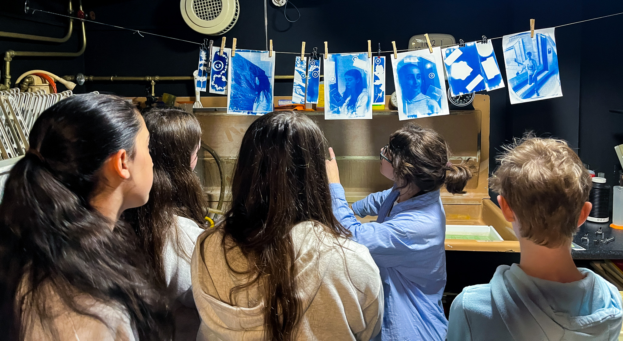 Young photographers create handmade cyanotype prints at Photo Museum Irelands Cruinniu na nOg workshop © Photo Museum Ireland2958 Alt text: A close-up of a table at an arts and crafts workshop where participants are creating collages. Various hands are seen working with materials, including a child using a glue stick and an adult drawing on paper. The table is covered with art supplies such as colored pencils, scissors, and various cut-out images, including photographs, artwork, and magazine clippings. The atmosphere appears creative and collaborative, with people of different ages participating in the activity.